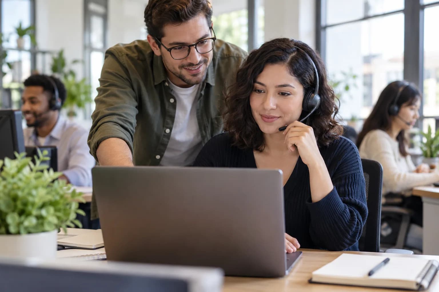 Customer support specialist wearing a headset at a laptop, colleague ready to help